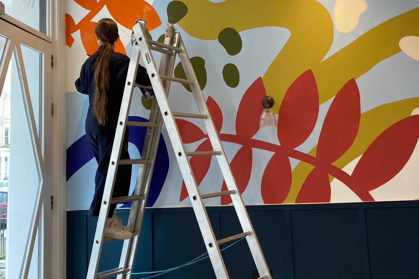 A decorator on a stepladder painting the bold abstract mural by hand, above dark blue panelling, with the sash window alongside