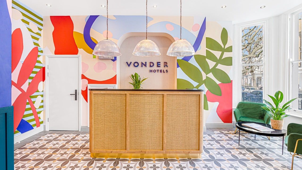 The finished aparthotel reception: a rattan-fronted desk on patterned encaustic floor tiles, with a full-height abstract mural in bright colours behind and glass pendant lights above