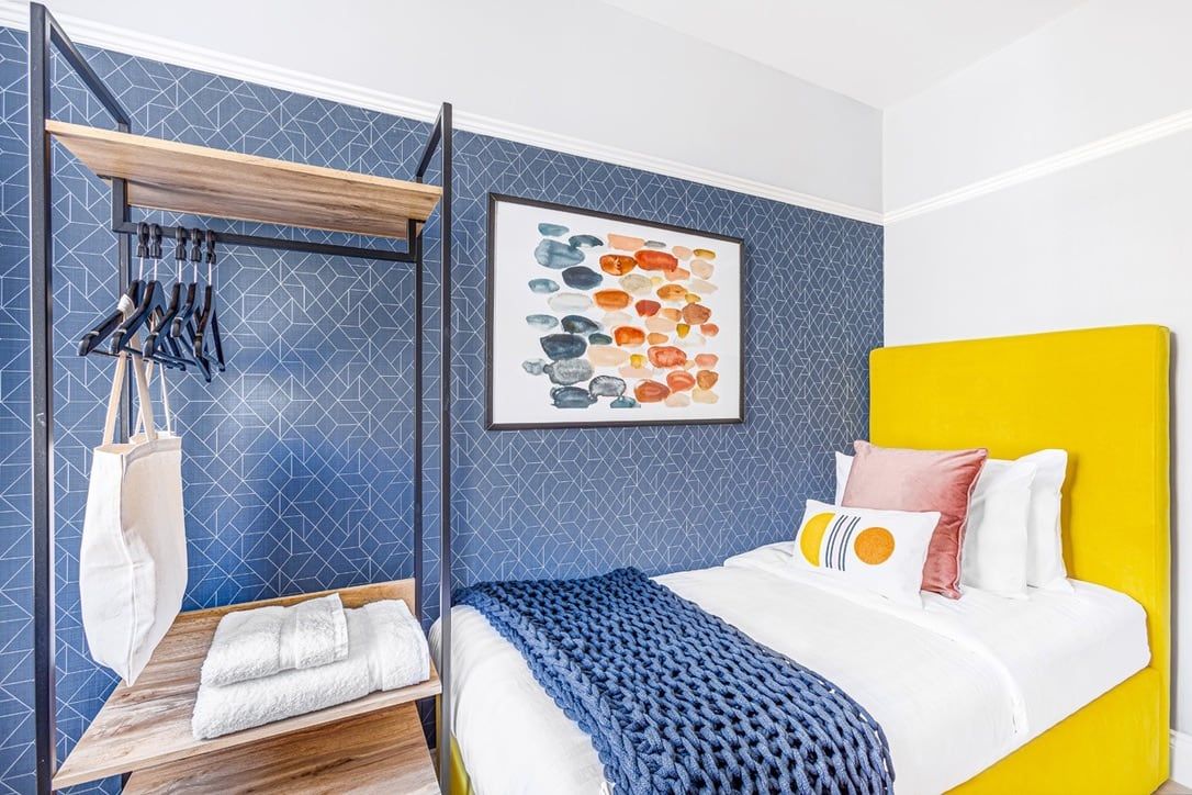 A finished bedroom with a blue panelled wall, yellow upholstered headboard, open hanging rail and framed artwork