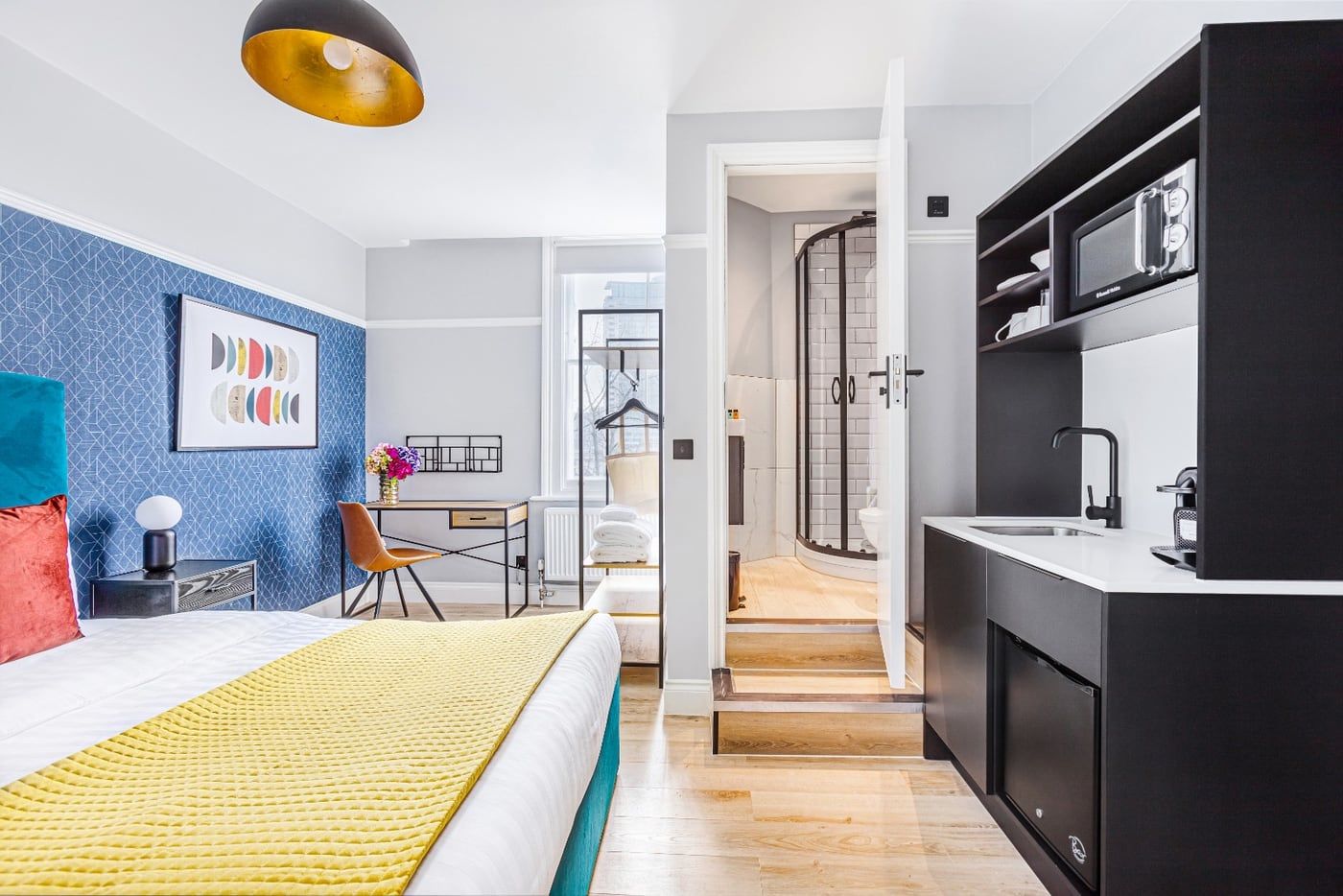 A finished studio: bed, desk and seating, with the fitted kitchenette and shower room visible through the opening