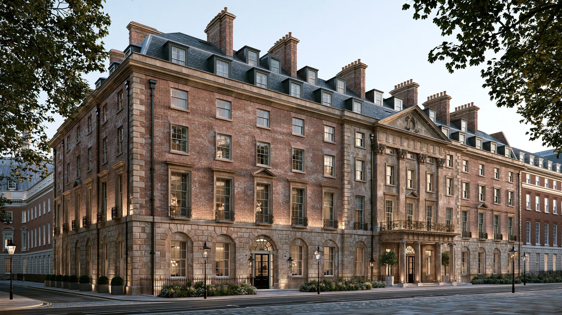 A Georgian terrace in red brick with stone dressings, a pedimented stone centrepiece and portico, iron balconies at first floor and a slate mansard roof lined with dormers and chimney stacks, shown at dusk