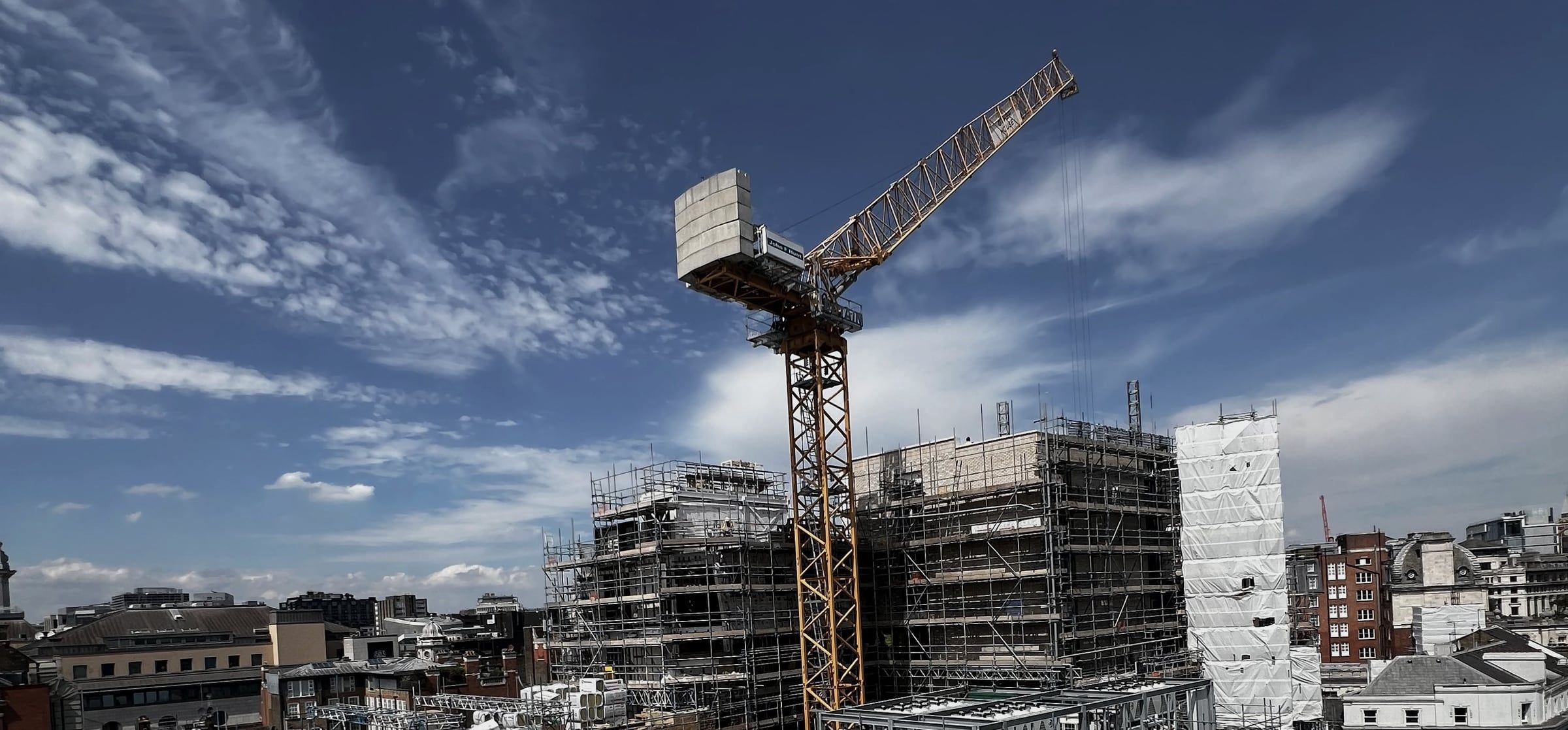 A tower crane above a mid-construction London building wrapped in scaffolding, period and modern rooftops beyond under a bright sky